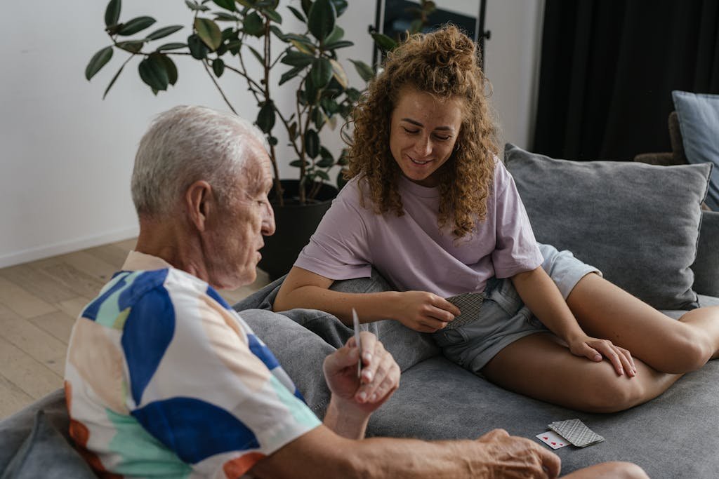 Un adulte senior et une jeune femme partagent un moment joyeux en jouant aux cartes sur un canapé confortable, à l’intérieur. Cette image a vocation à montrer la transmission entre les vieilles et nouvelles générations.